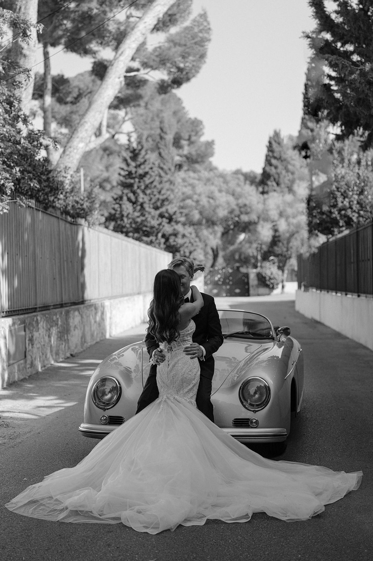 Couple kisses leaning on vintage Porsche convertible with lace mermaid train fanned across road in B&W
