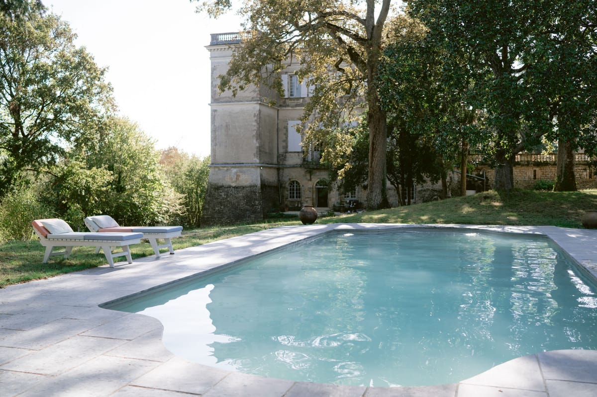 Limestone chateau with white shutters and balustraded roofline behind rectangular swimming pool and sun loungers