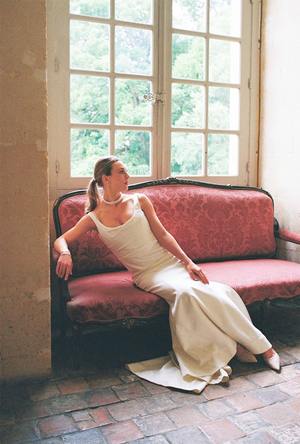 Analog frame of bride reclining on red damask sofa by tall French windows