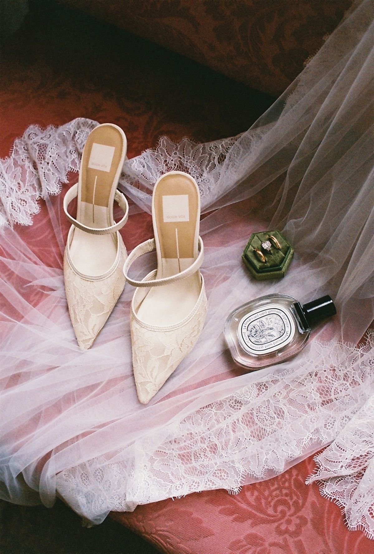 Film flat lay of lace bridal heels, ring box and perfume on tulle veil and damask cushion