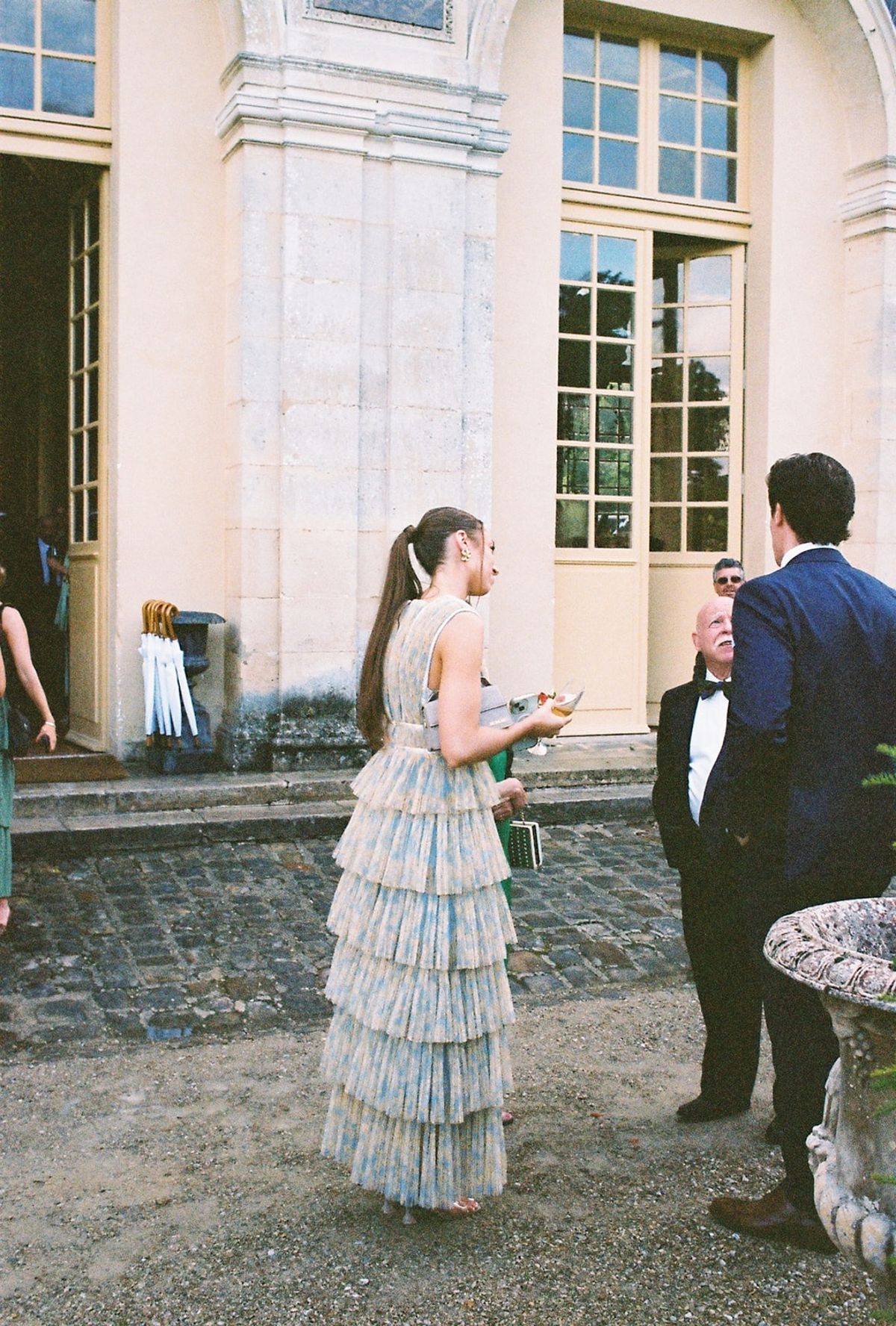 Guest in tiered pastel gown chats outside lit chateau pavilion at twilight
