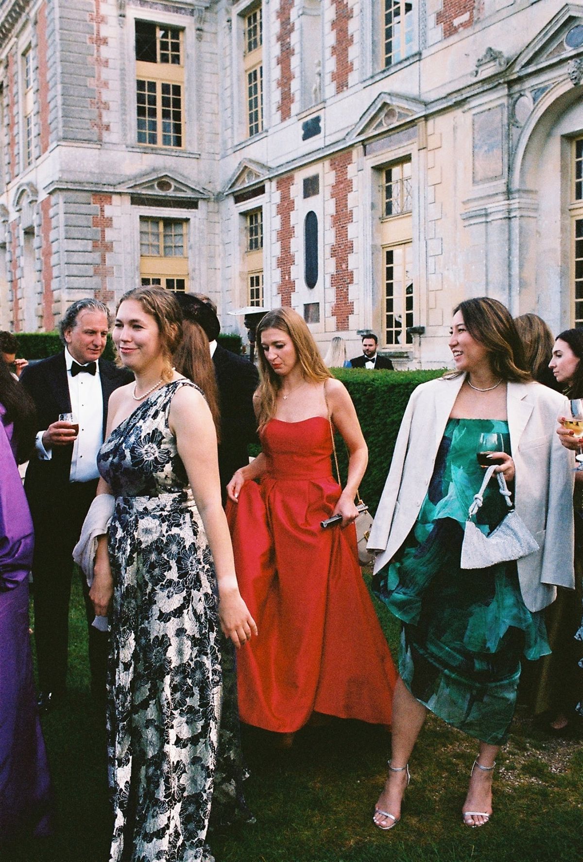 Guests in floral and jewel-toned gowns move across chateau lawn during cocktail hour