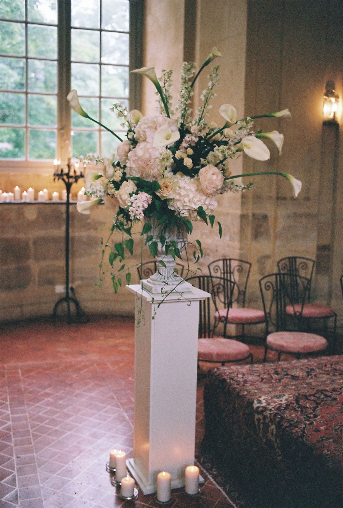 Tall ivory and blush calla lily arrangement on white pedestal beside iron candelabra