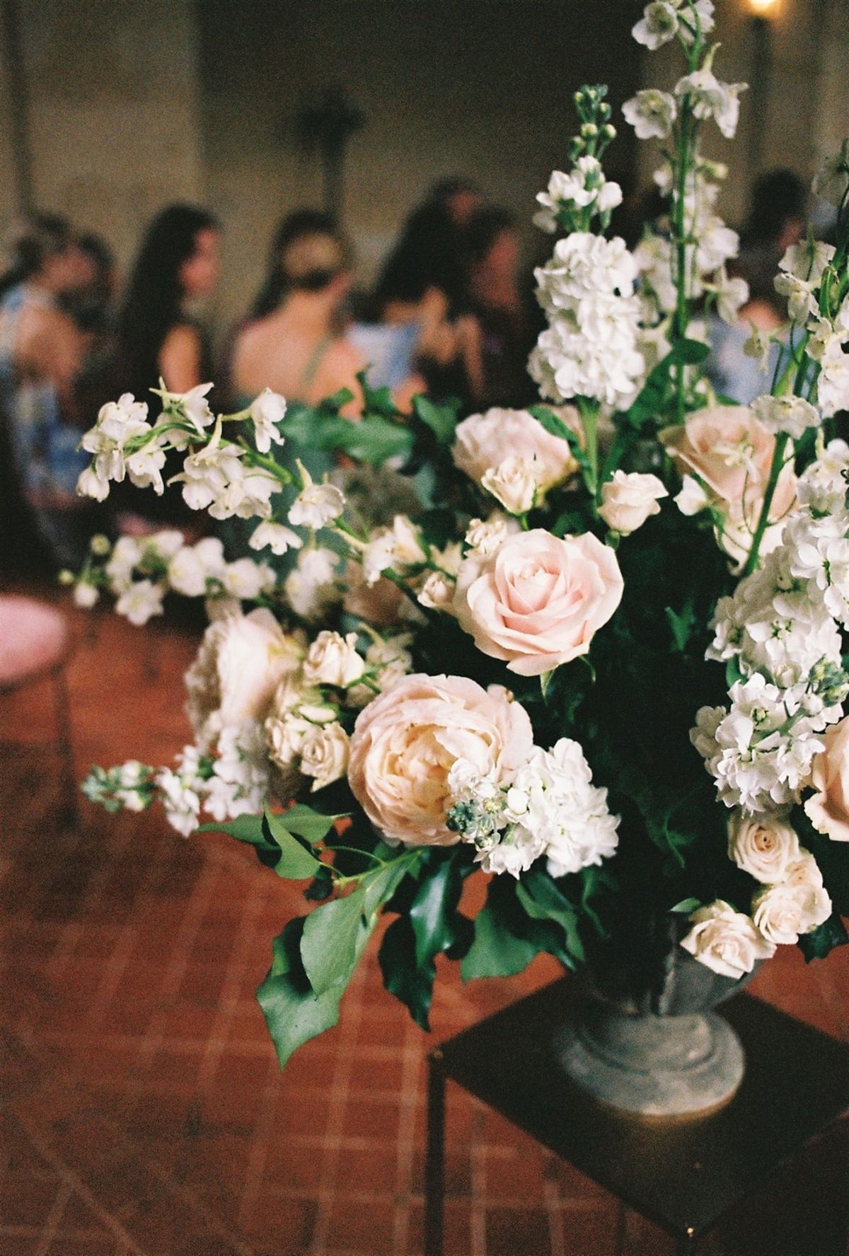Close ceremony detail of blush and ivory urn arrangement with guests softly behind