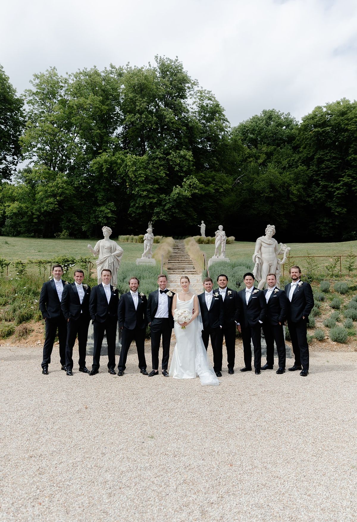 Bride and groom stand with groomsmen on gravel beneath lavender staircase with statues