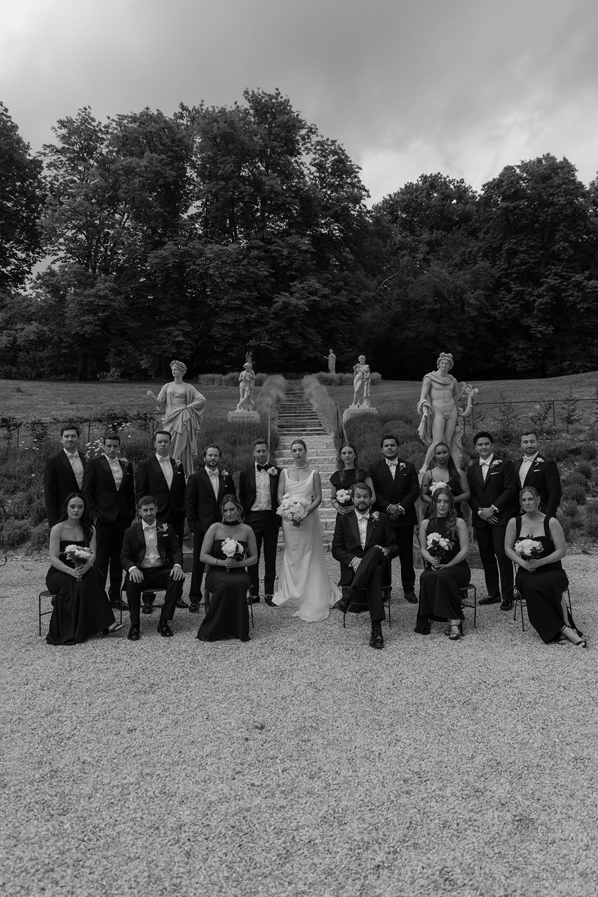 Full bridal party in two rows under lavender garden staircase with statues behind