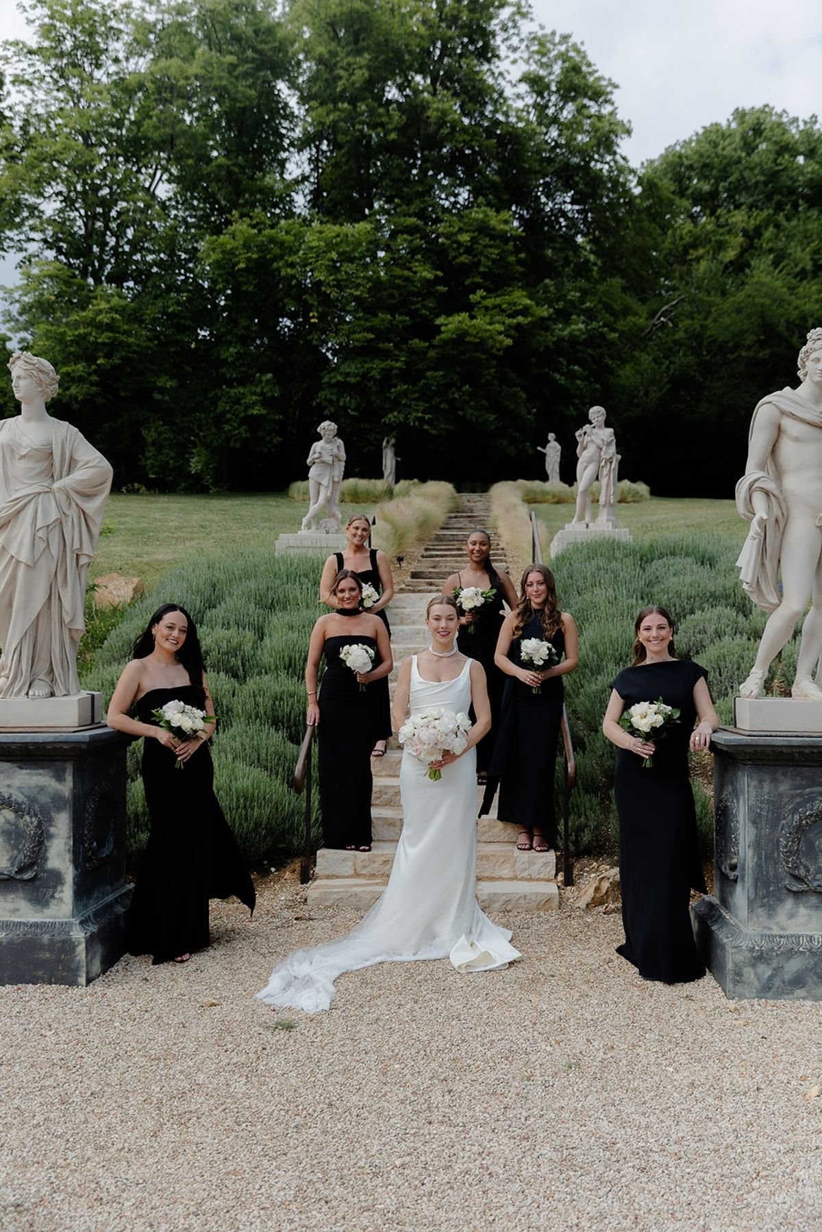 Bride and bridesmaids in black gowns at foot of lavender garden steps with bouquets
