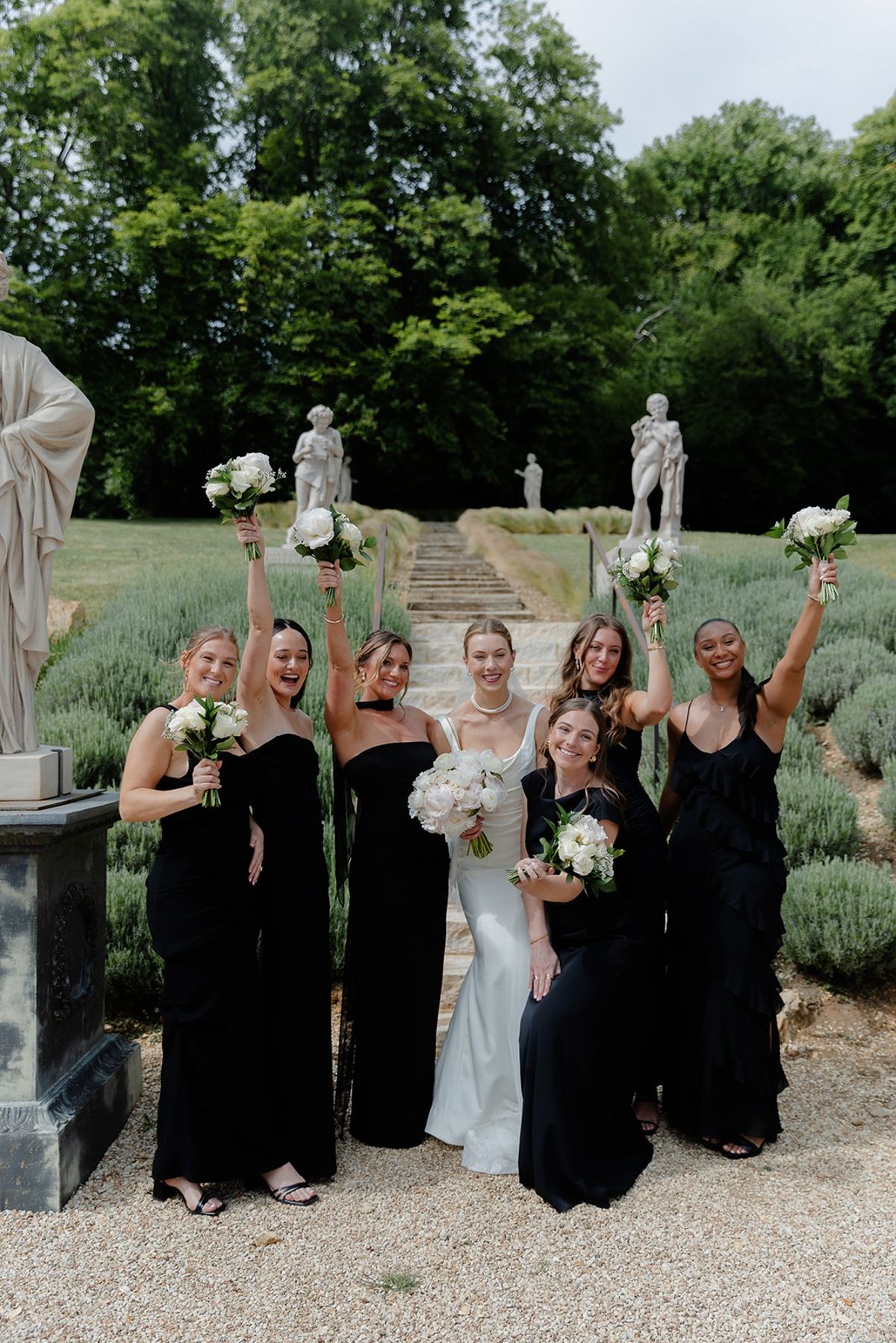 Bride and bridesmaids raise bouquets overhead on lavender garden steps