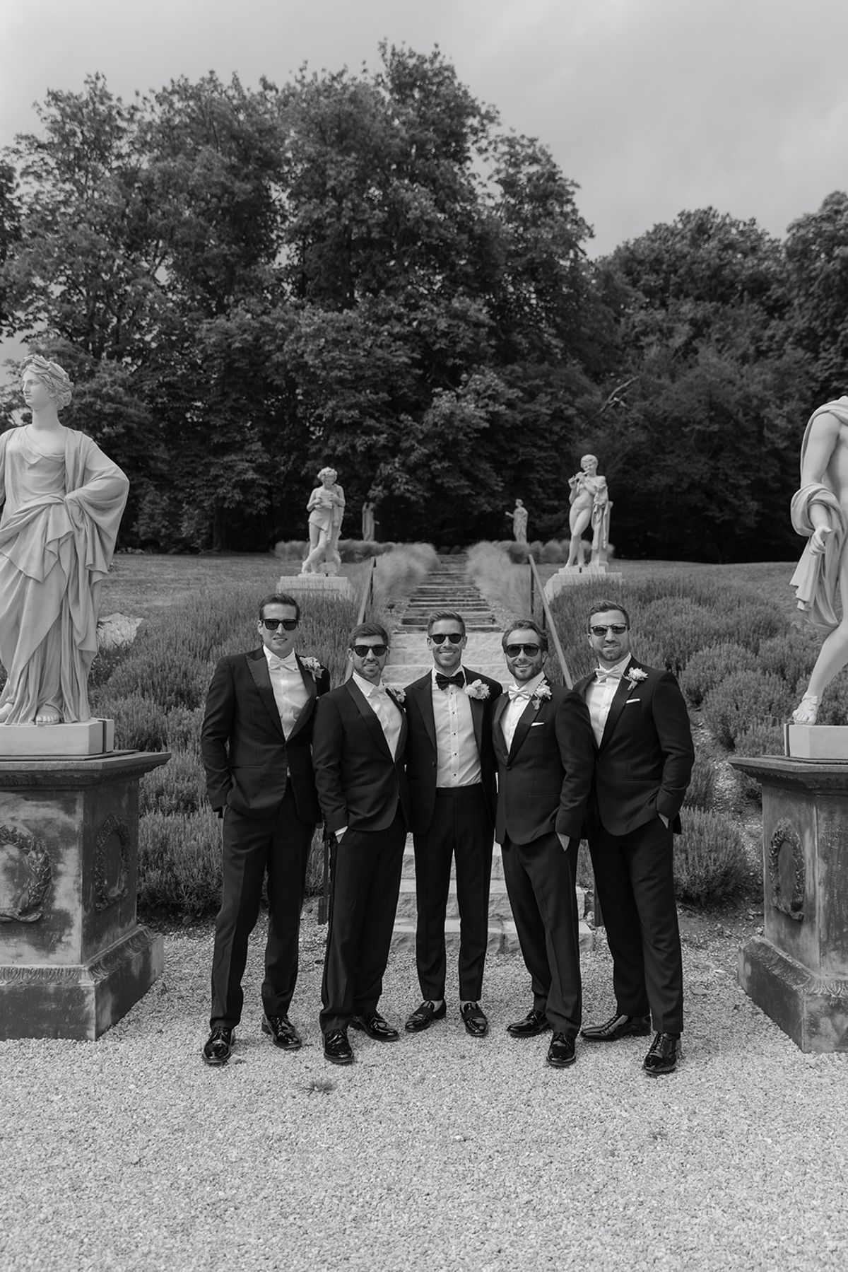 Groom and groomsmen in tuxedos at base of lavender staircase with statues