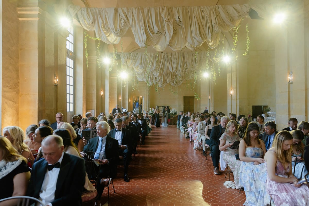 Guests fill long ceremony hall under sculpted fabric canopy with trailing greenery
