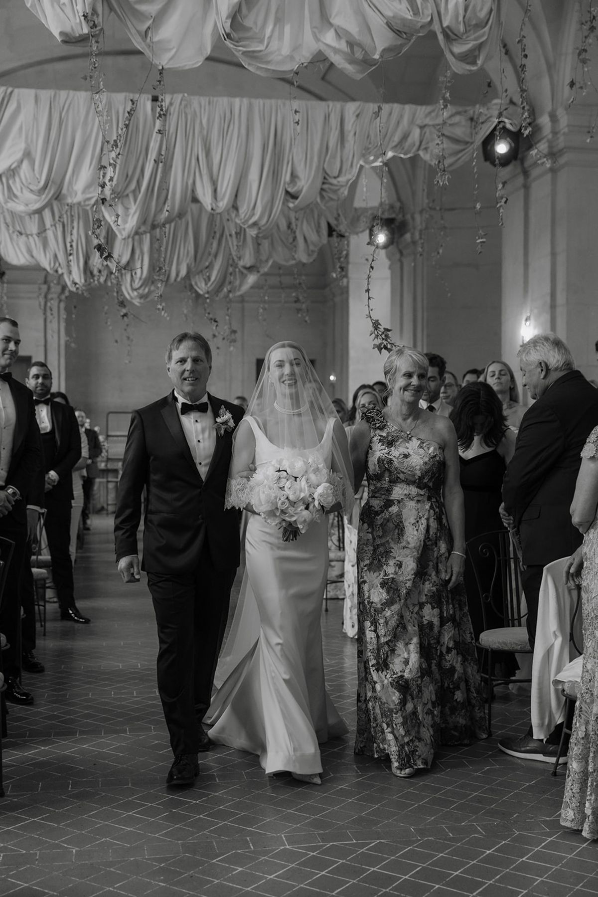 Bride processes down aisle between parents under draped fabric ceiling