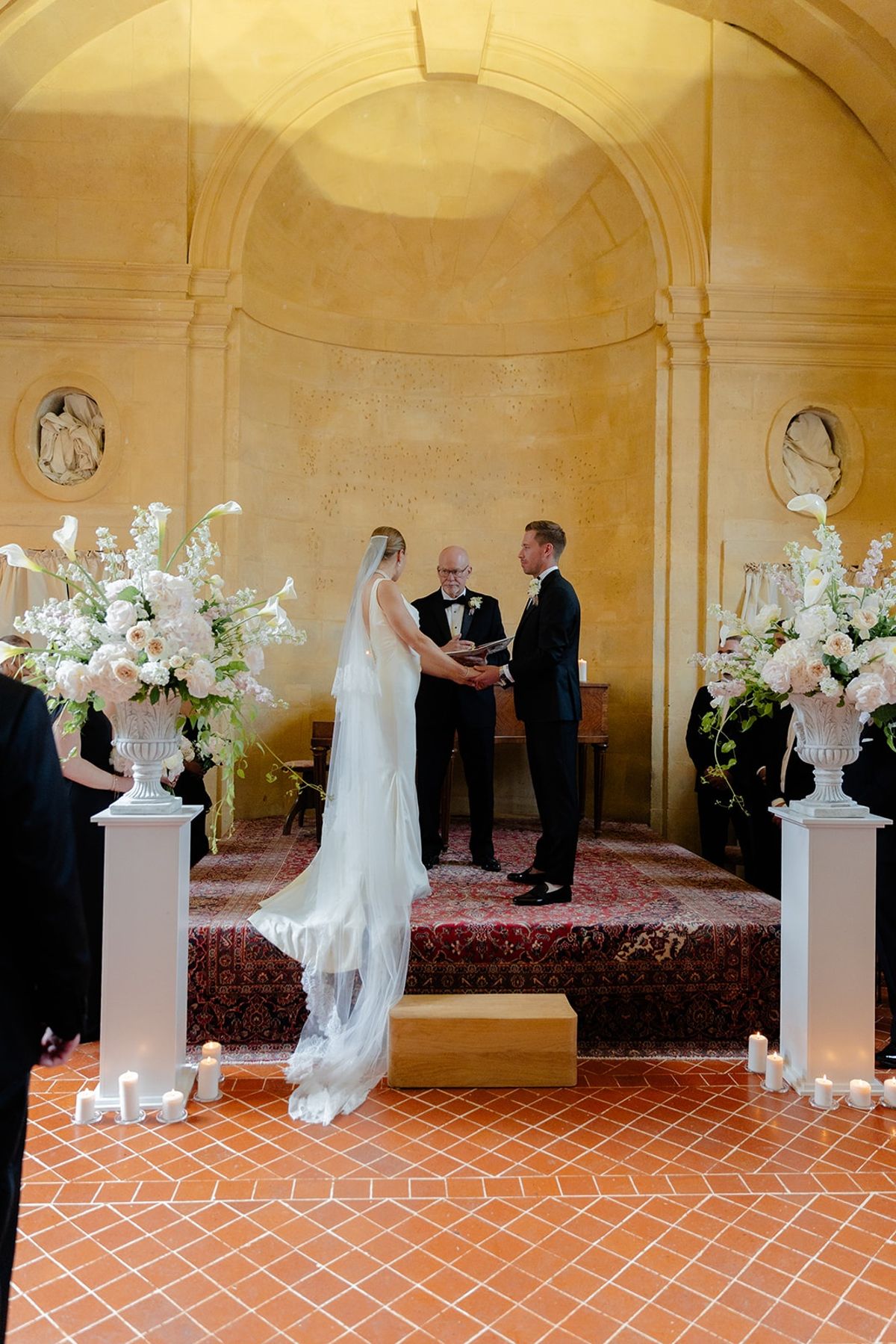 Couple holds hands at altar between tall blush and white floral urns
