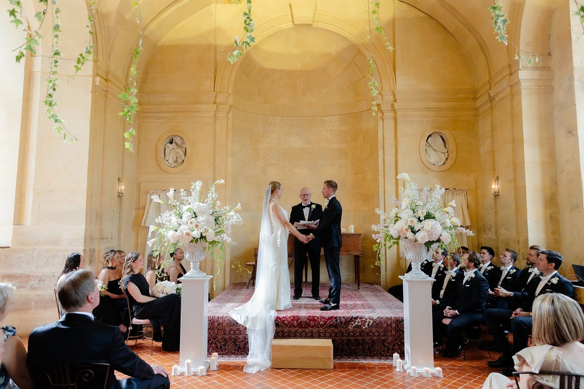 Wide ceremony view with couple holding hands between blush floral urns and guests