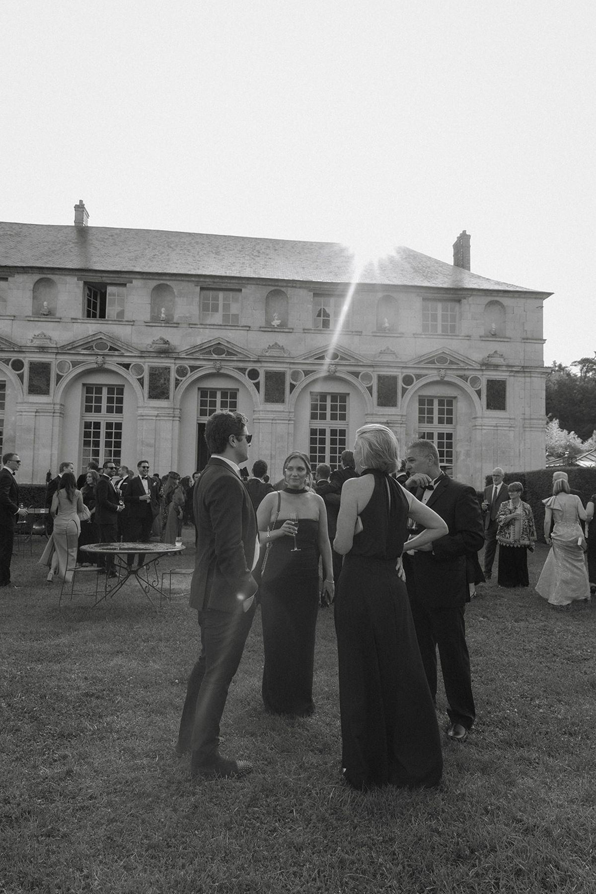 Guests in formal dress gather on chateau lawn after ceremony with stone facade behind