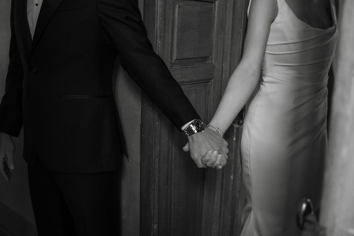 Bride and groom clasp hands at centre with watch and bracelet visible by door