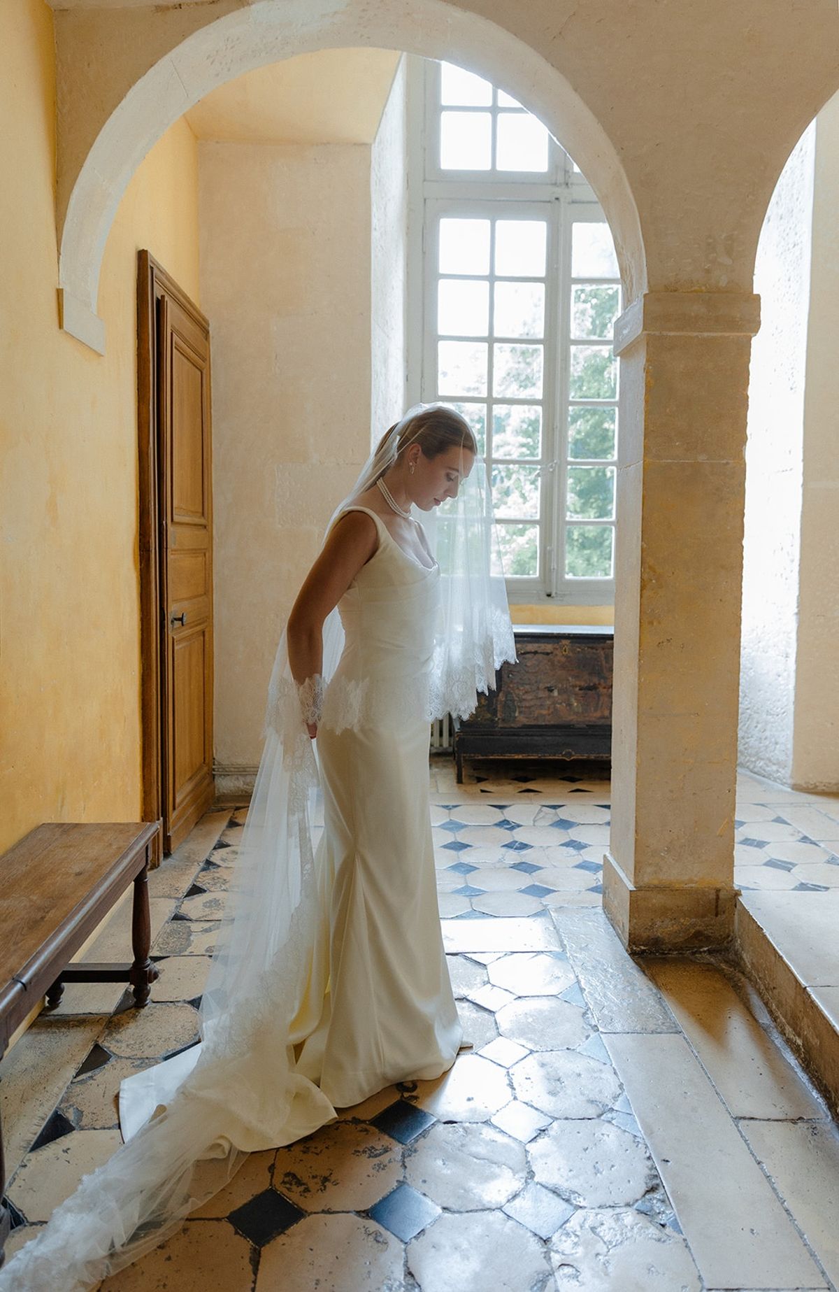 Bride in yellow stone arcade holds lace veil edge under tall arched window