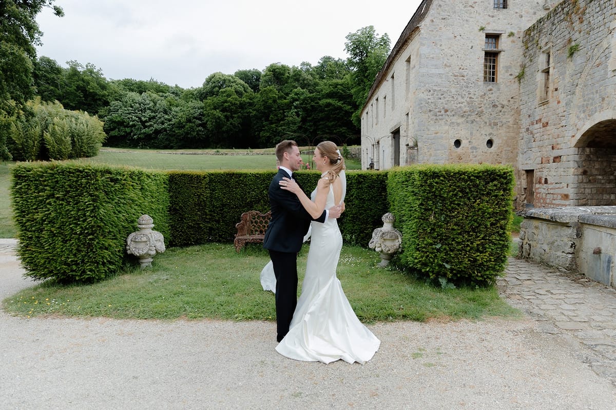 Bride and groom embrace on gravel path between hedges with chateau outbuilding behind