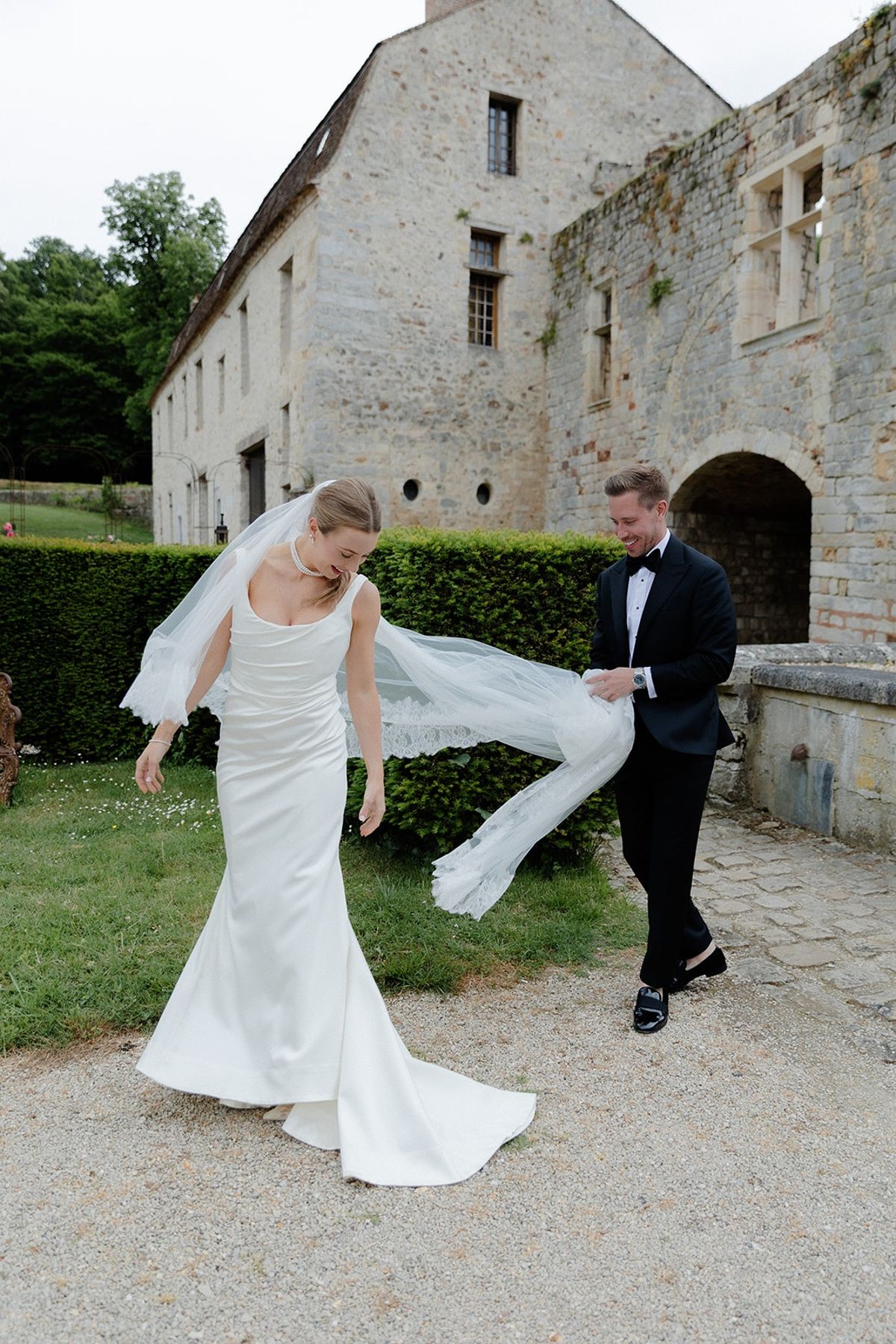 Groom lifts trailing lace veil for bride as they walk along cobbled chateau path