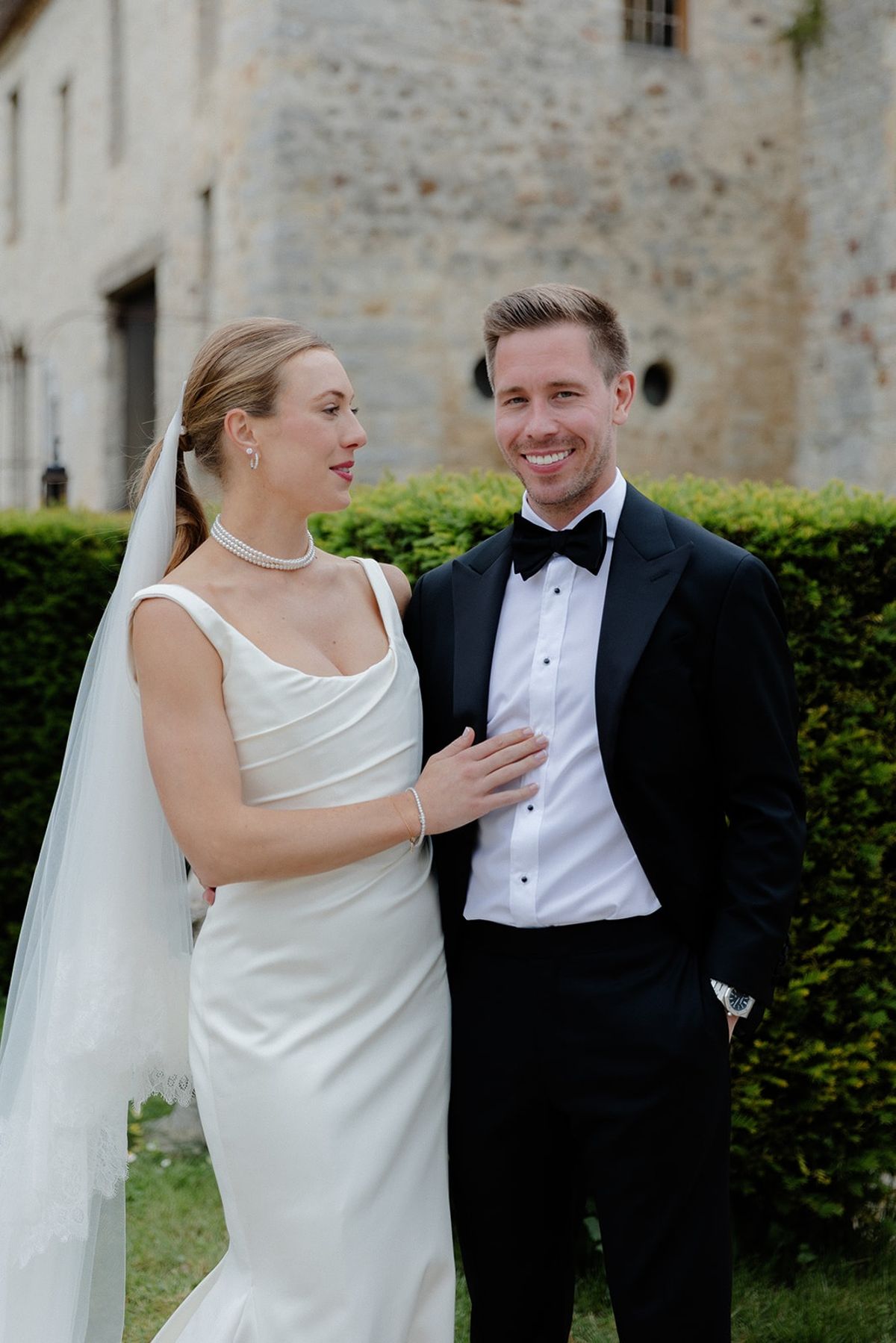 Bride rests hand on groom's chest as he smiles at her by chateau hedge