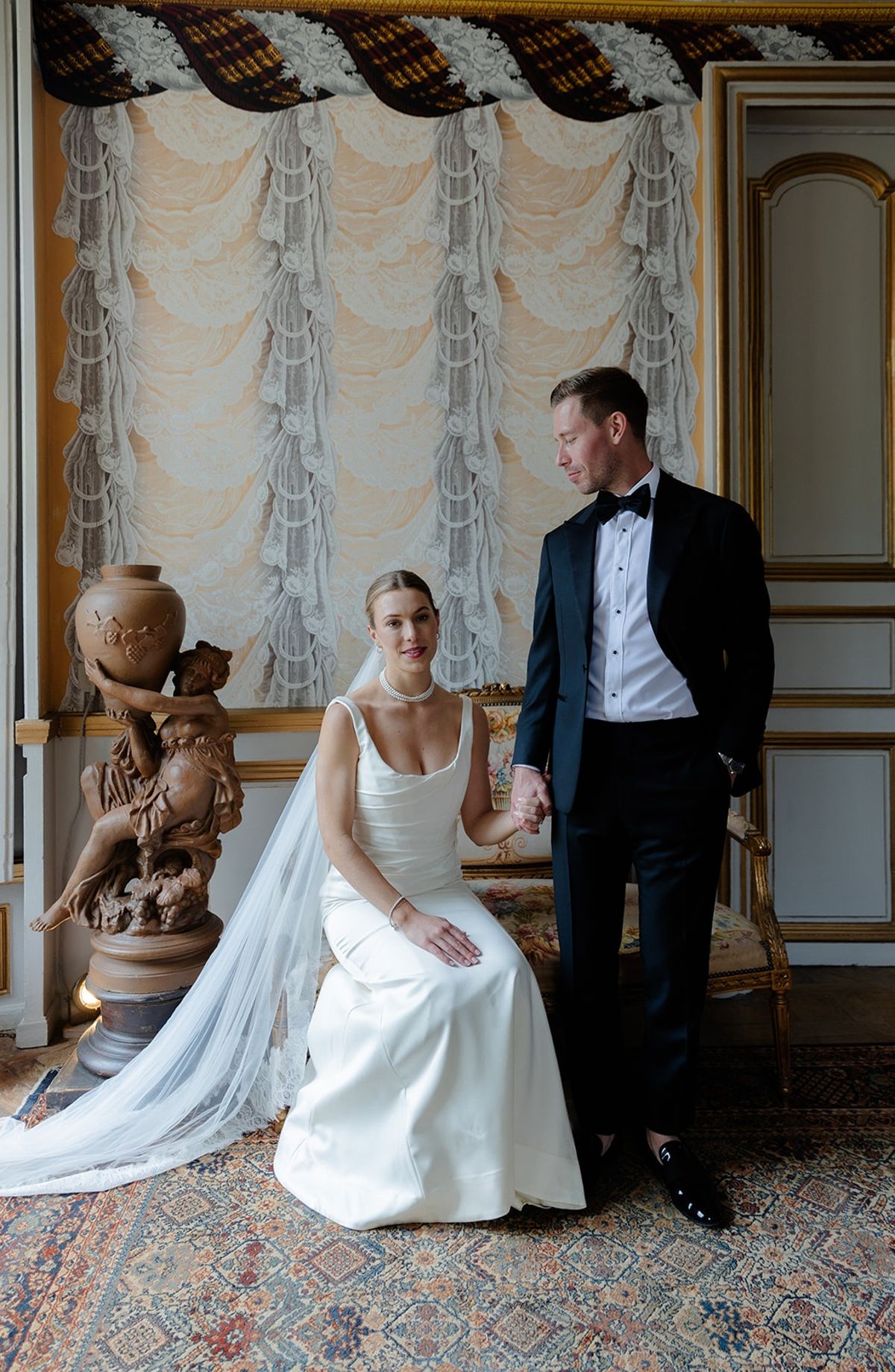 Bride sits on gilt sofa beside standing groom in front of antique lace salon backdrop