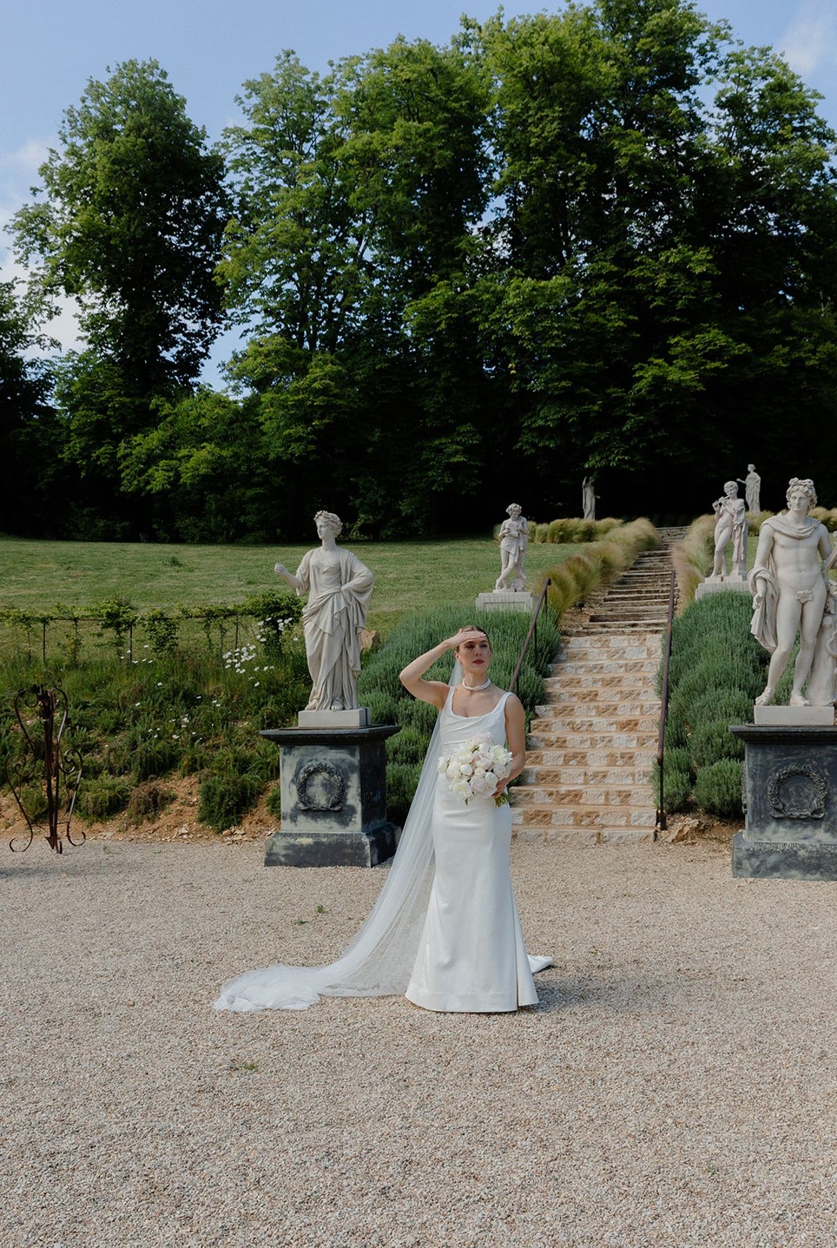 Bride holds bouquet by lavender staircase between marble statues in chateau garden