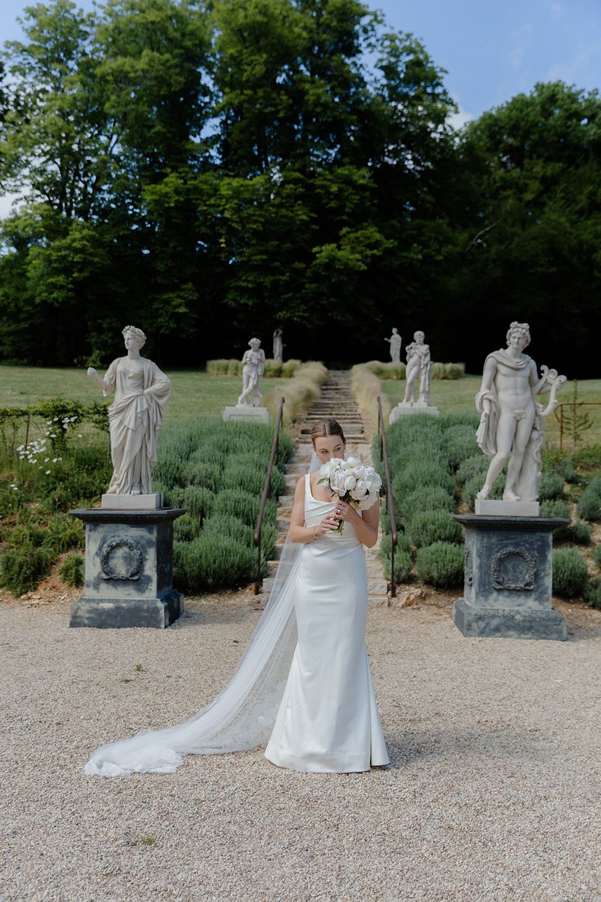 Bride lifts bouquet to her face at foot of lavender allee between classical statues