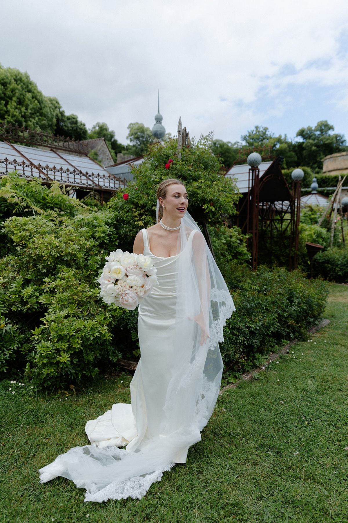 Bride holds bouquet of peonies and roses in chateau kitchen garden by conservatory
