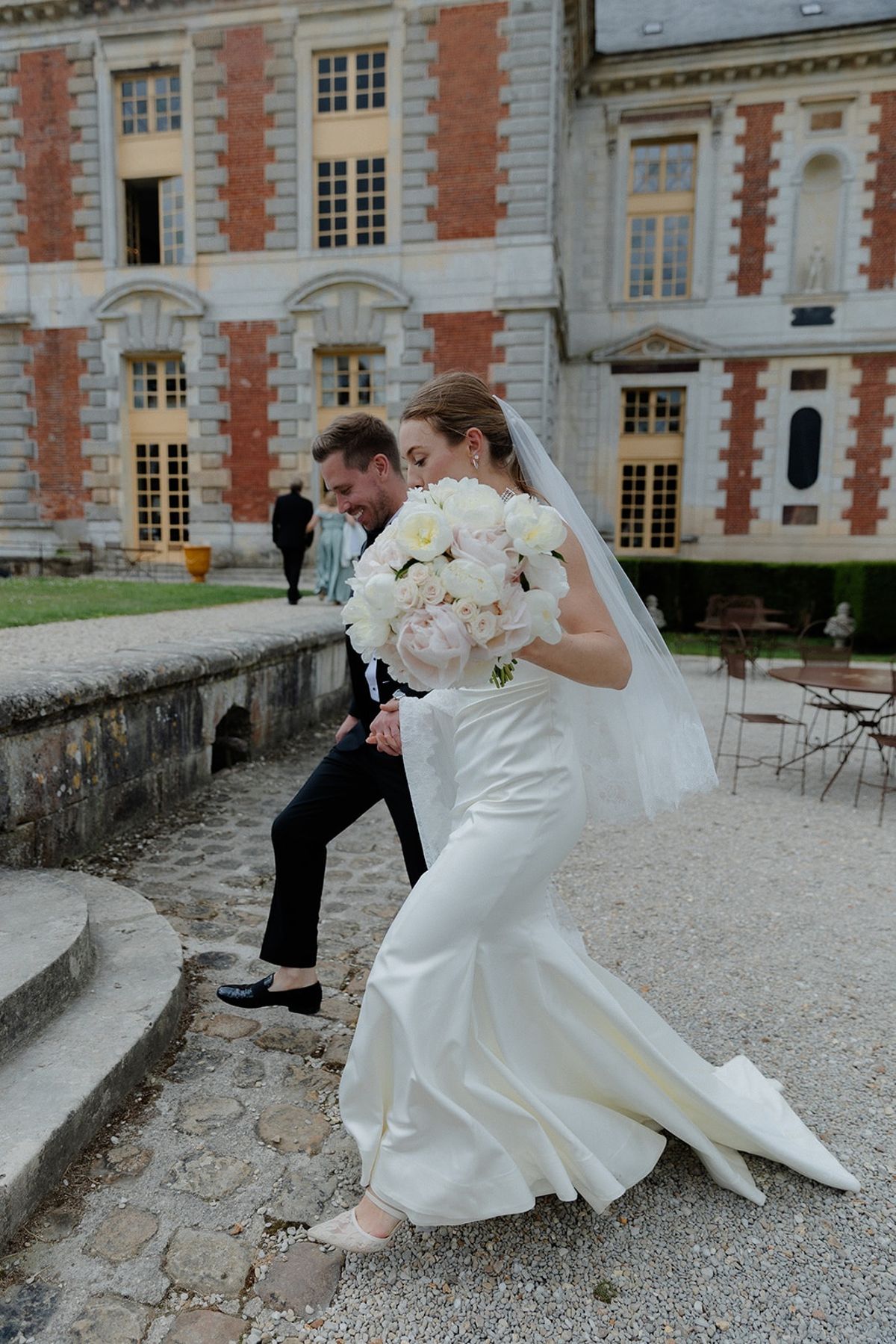 Bride leads groom across chateau forecourt holding bouquet aloft as both laugh