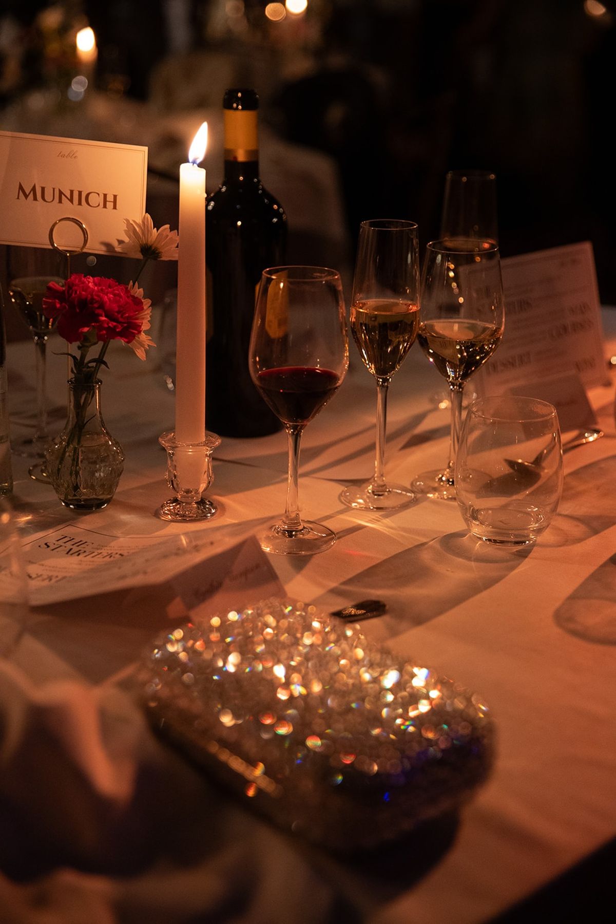 Candlelit dinner detail with Munich table card, rose bud vase, wine and beaded clutch