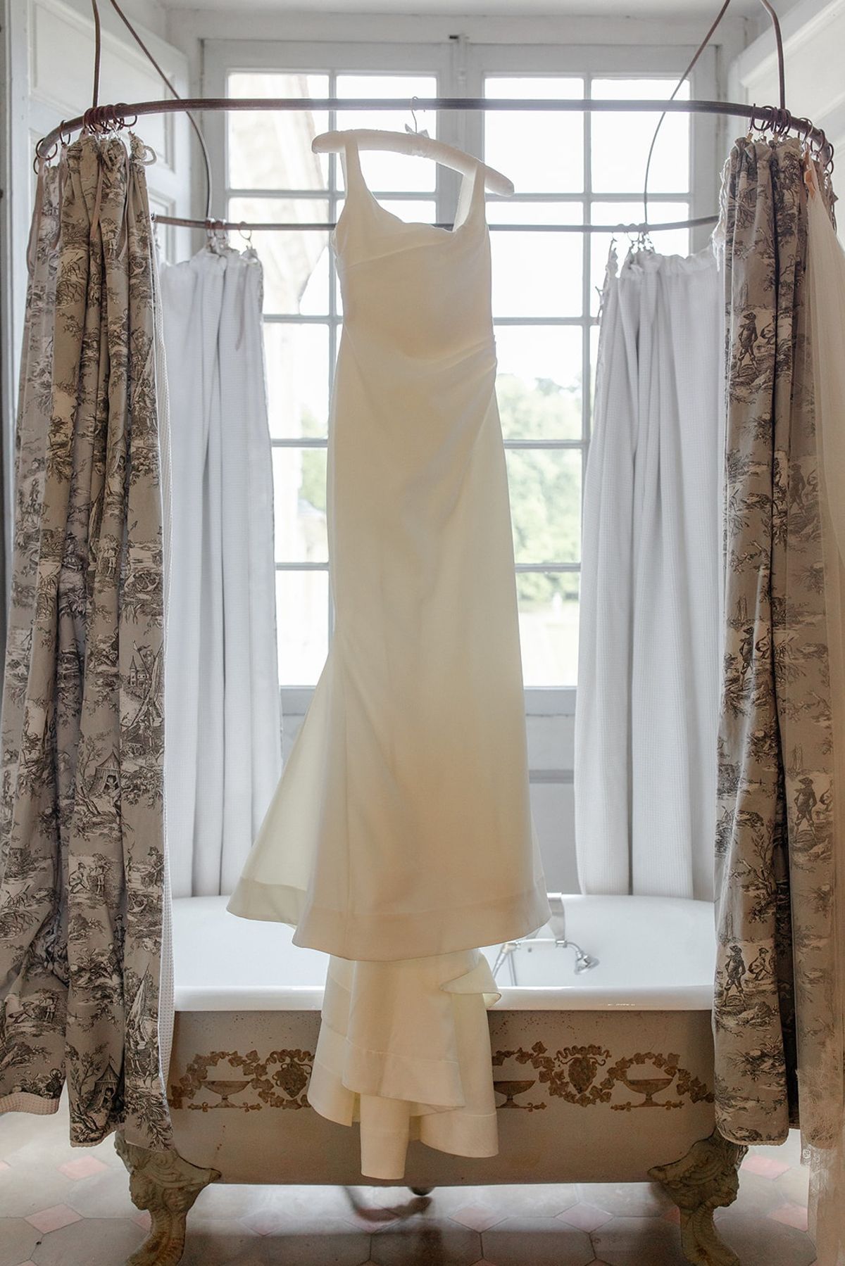 Bridal gown hangs above ornate bathtub between toile de Jouy curtains at a chateau