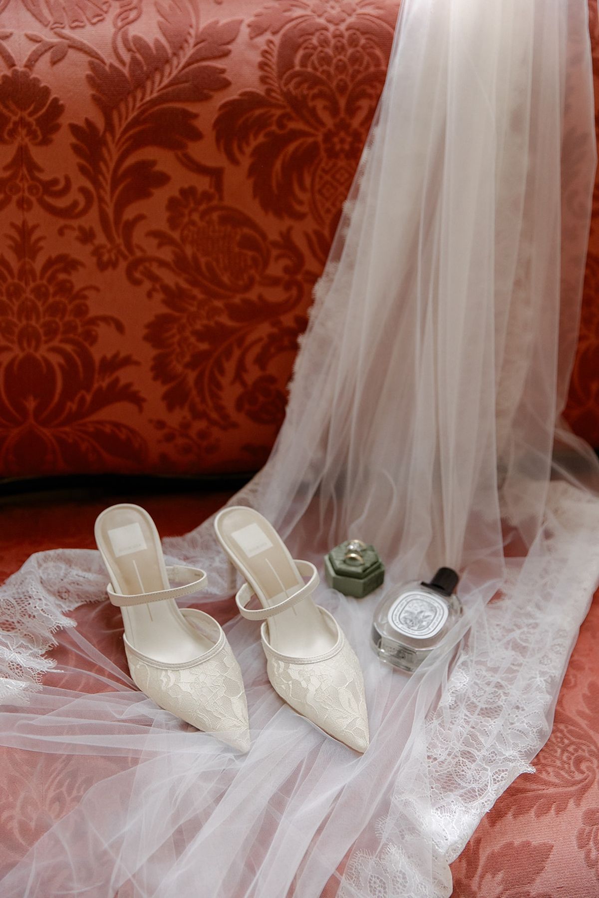 Bridal heels, perfume and ring box arranged on lace veil over damask cushion