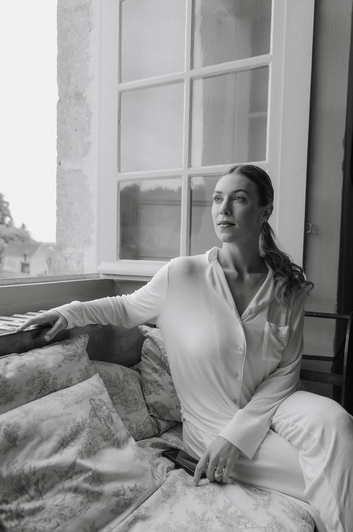 Bride sits by tall windows in matching pyjamas resting against toile cushions