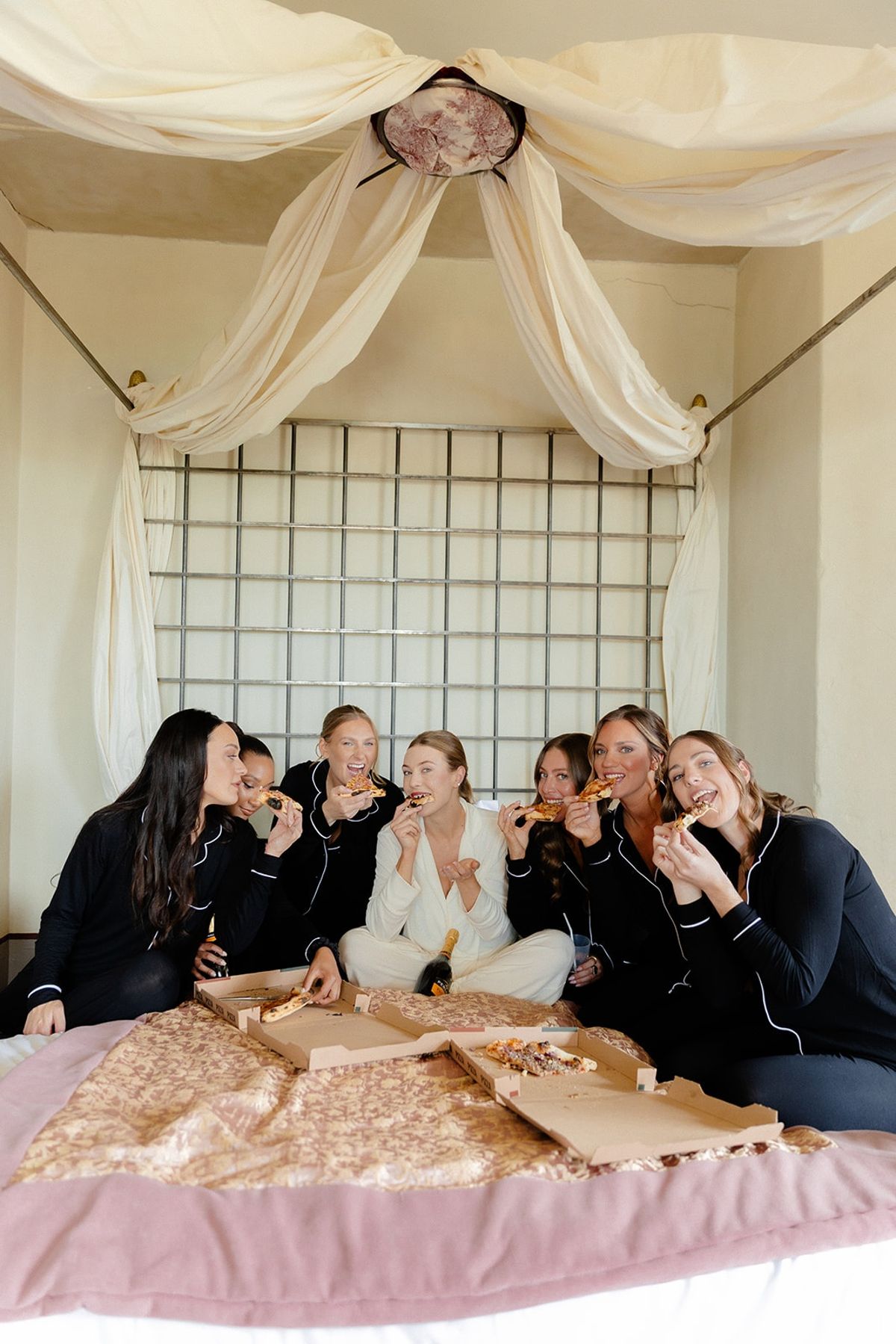 Bride and bridesmaids share pizza on a canopied bed in matching pyjamas