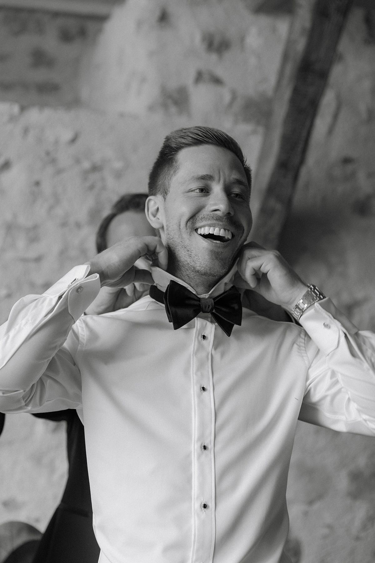 Groom laughs while adjusting bow tie against rough stone chateau wall