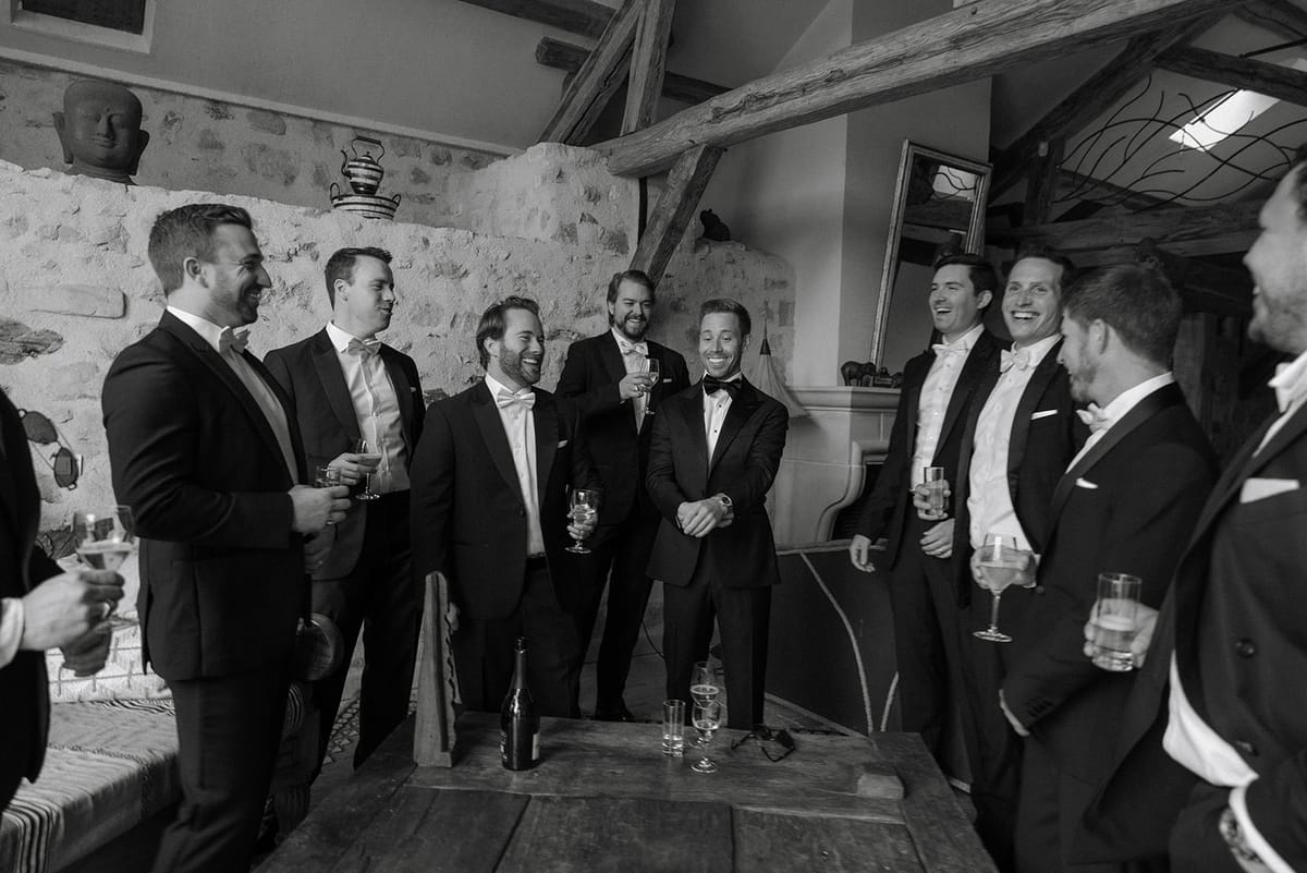 Groom and groomsmen in tuxedos raise glasses around a rustic wooden table
