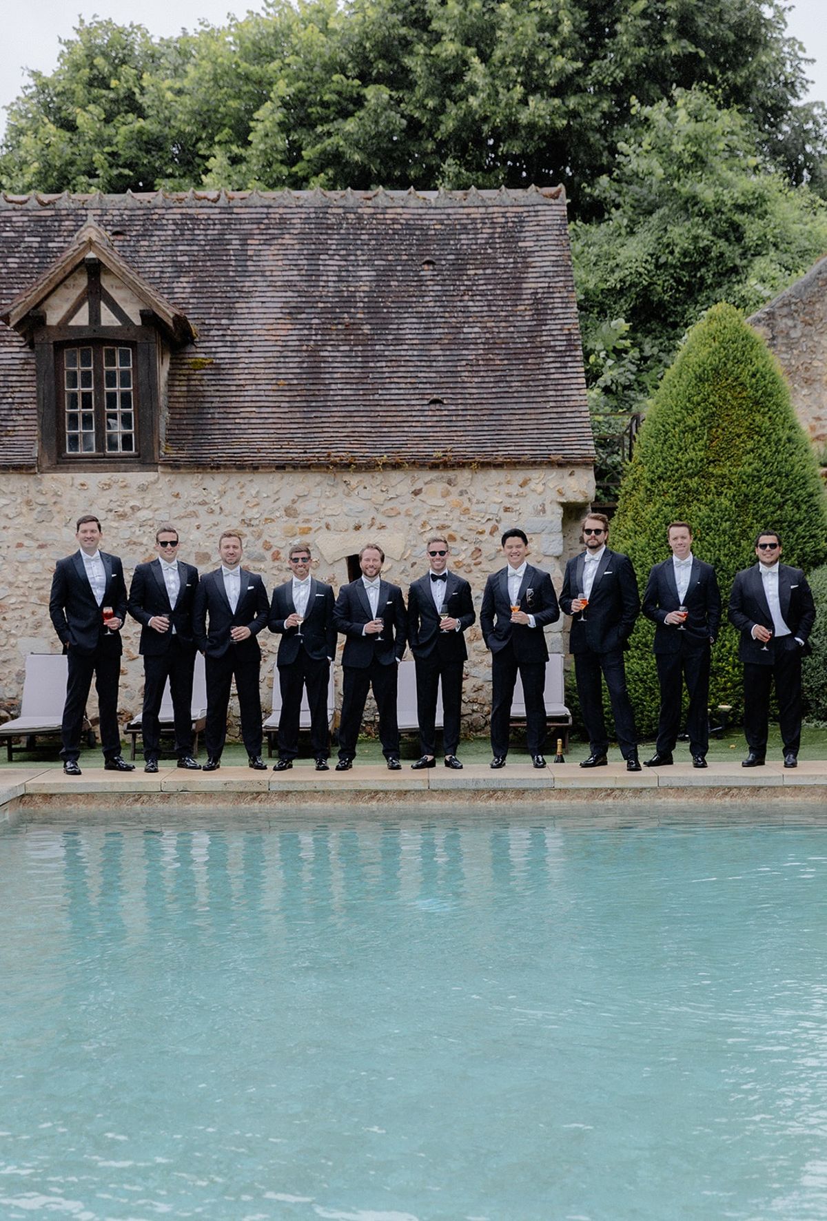 Groomsmen line up along chateau pool in matching tuxedos with sunglasses