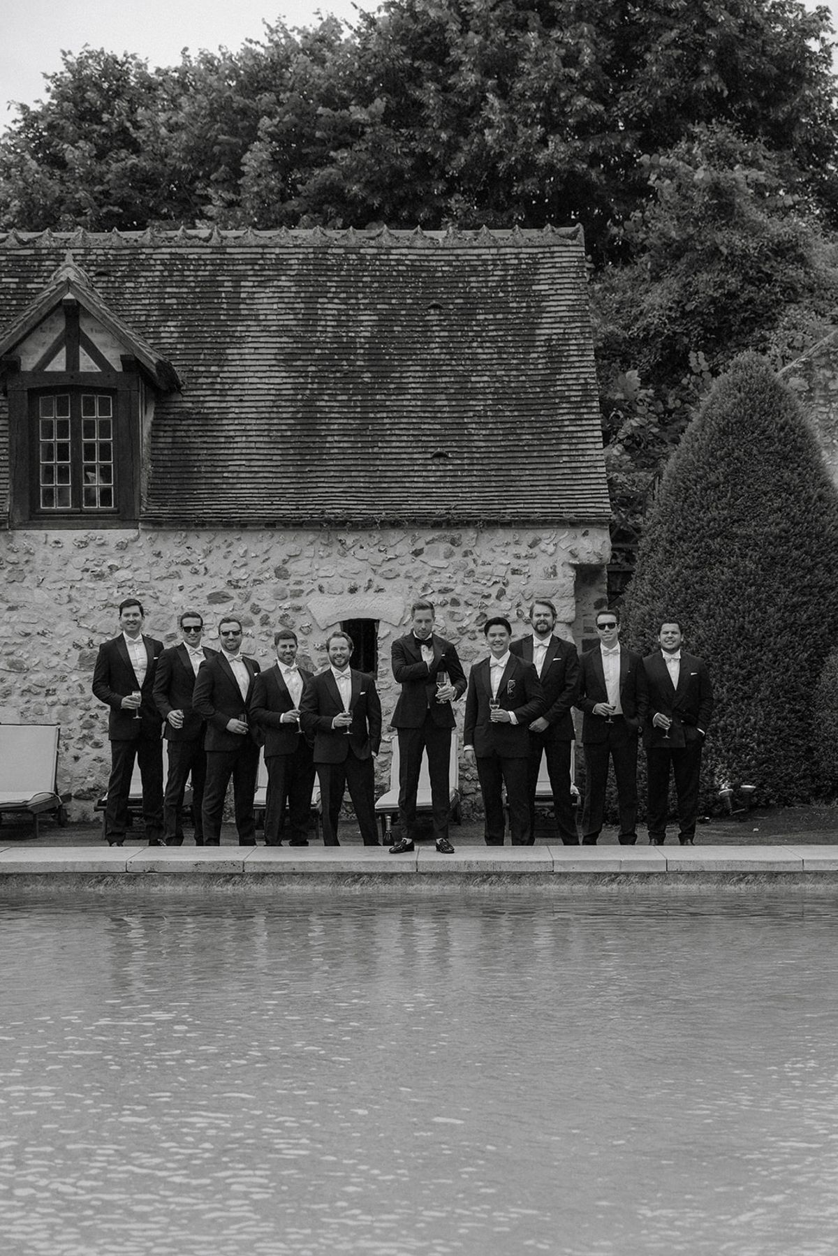 Groomsmen pose along chateau pool with half-timbered outbuilding behind