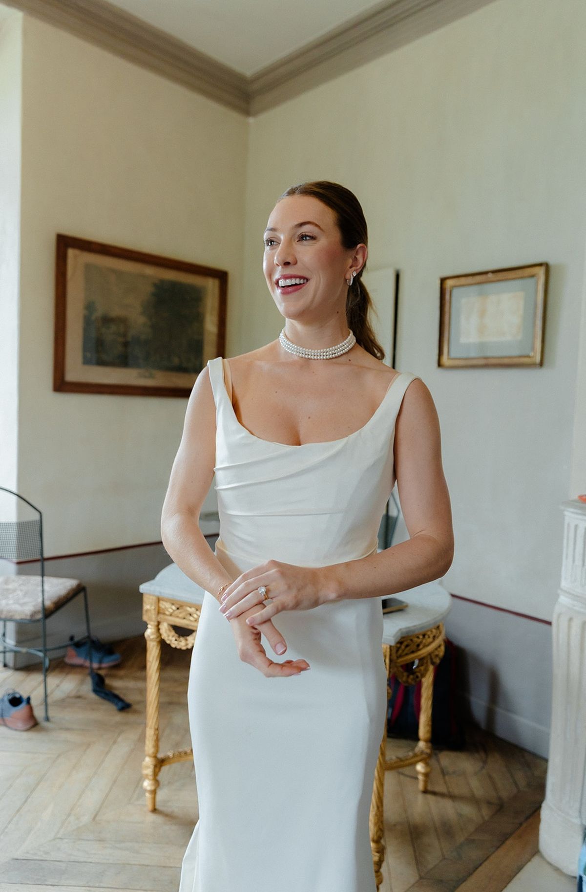 Bride in satin dress and pearl choker stands in a pale chateau drawing room