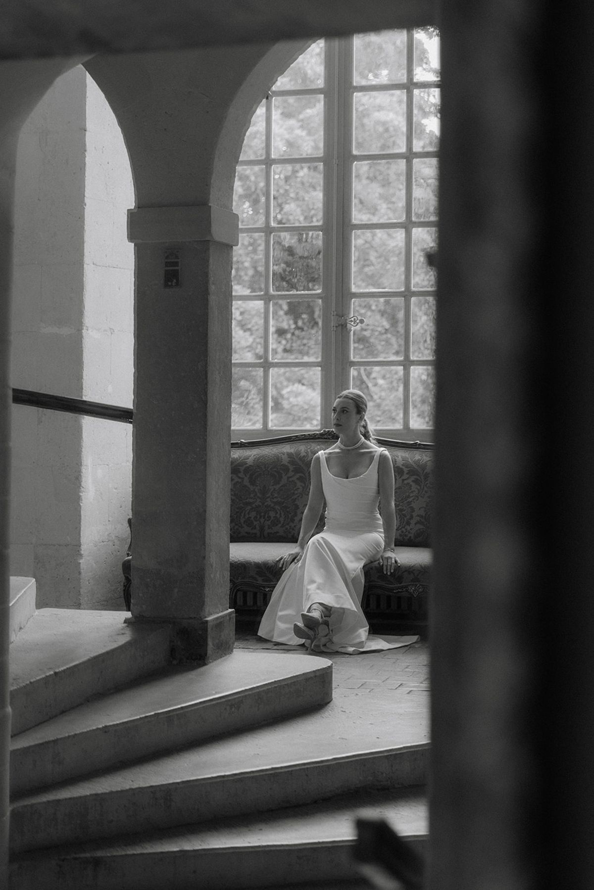 Bride sits on damask settee in chateau stone stairwell beneath tall arched window