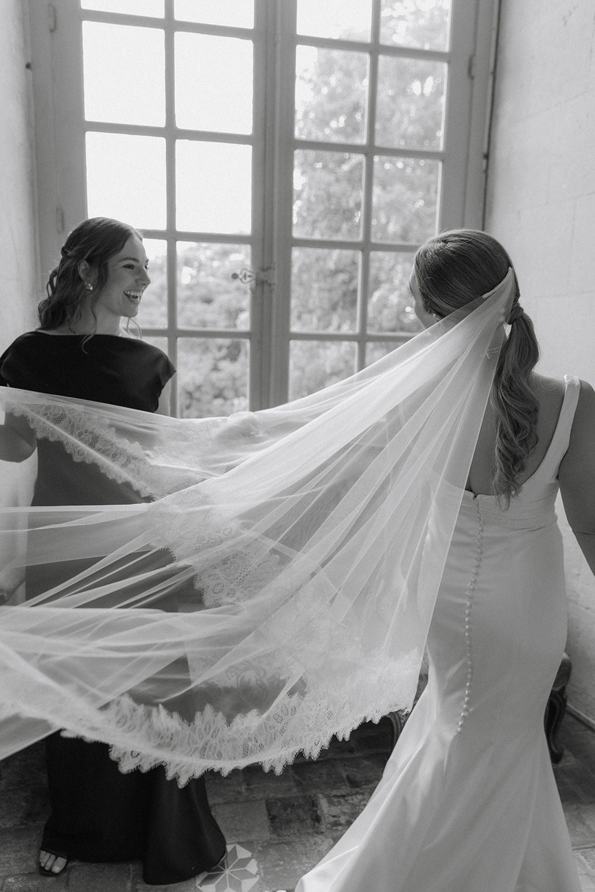 Bridesmaid lifts the cathedral lace veil as bride turns toward chateau window