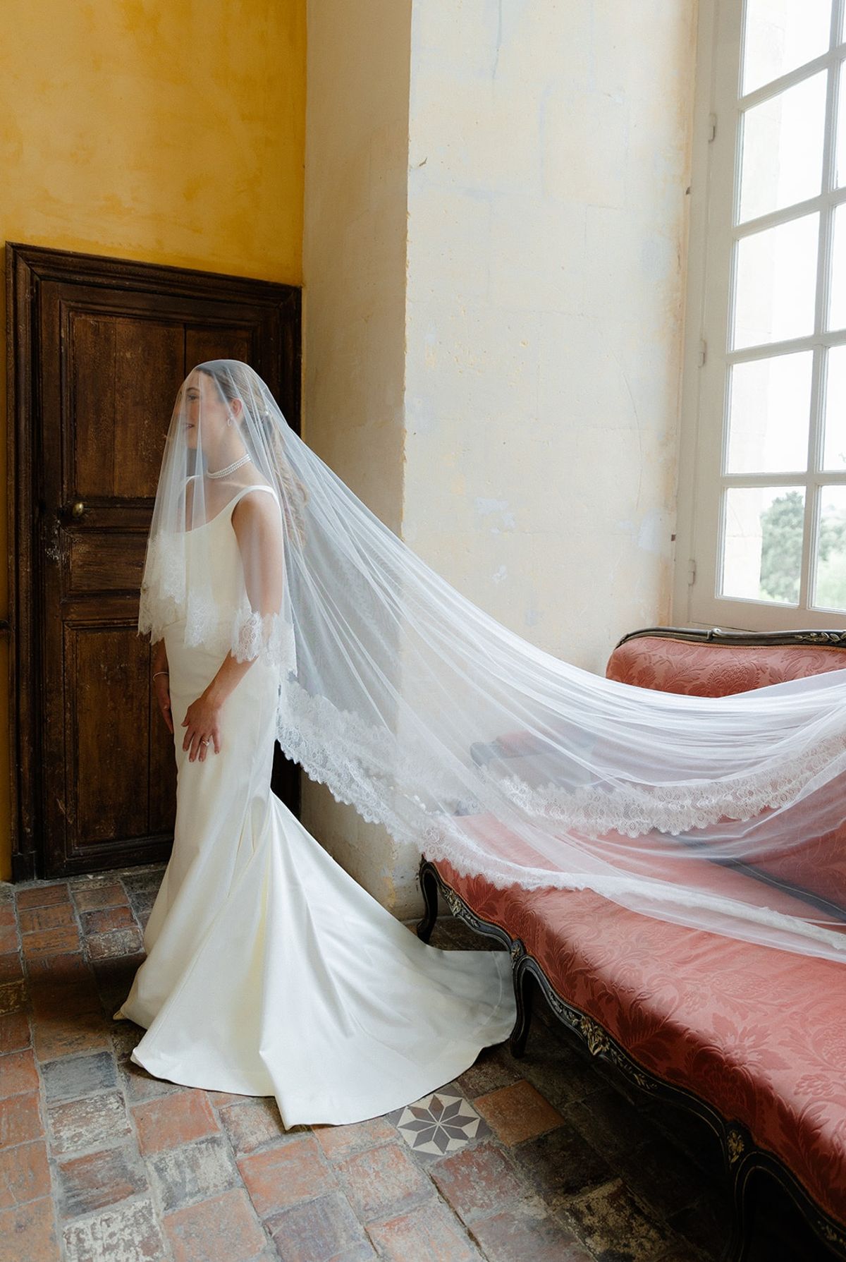 Bride poses with cathedral lace veil pulled over face beside ornate sofa