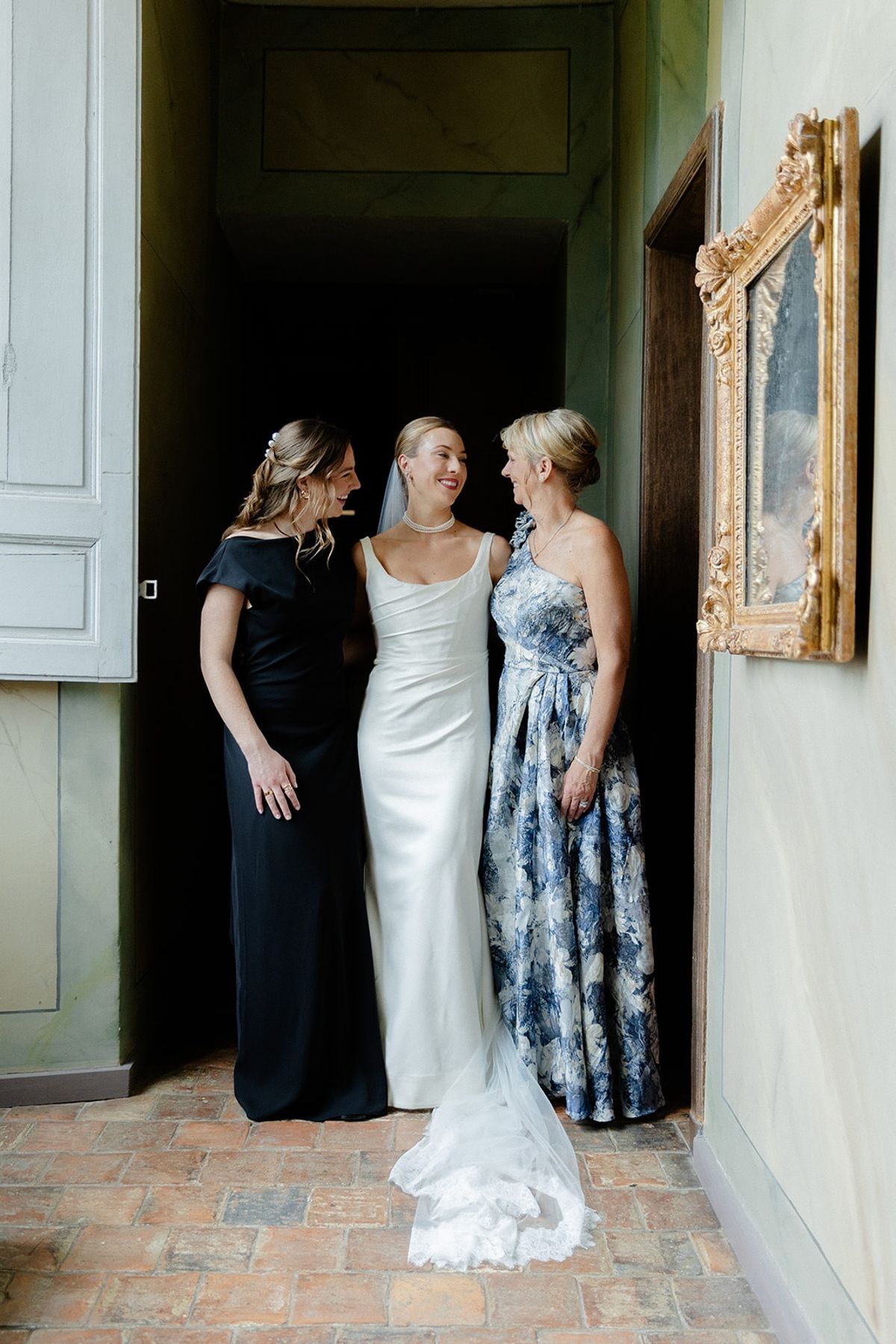 Bride with mother and bridesmaid pause in green marbled chateau corridor