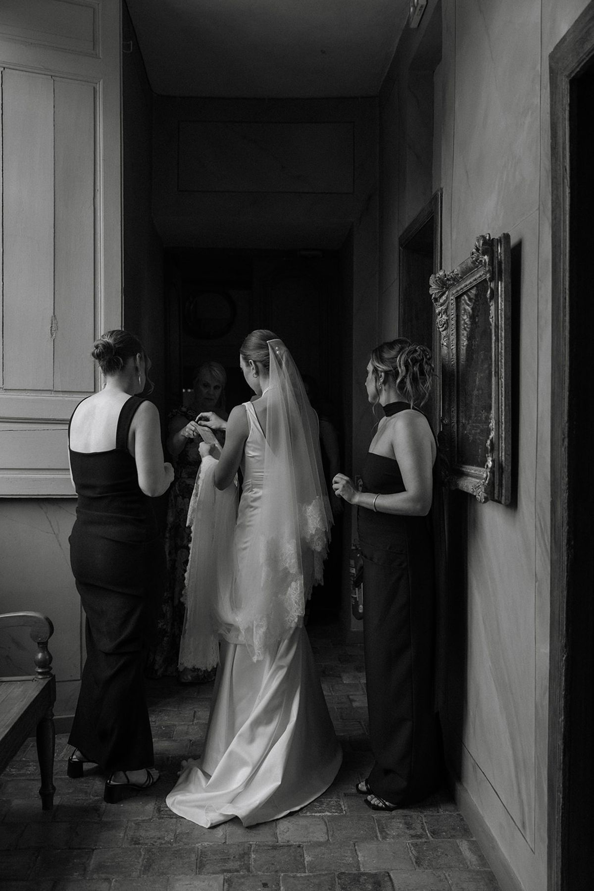Bridesmaids and mother gather around bride to adjust veil in chateau corridor