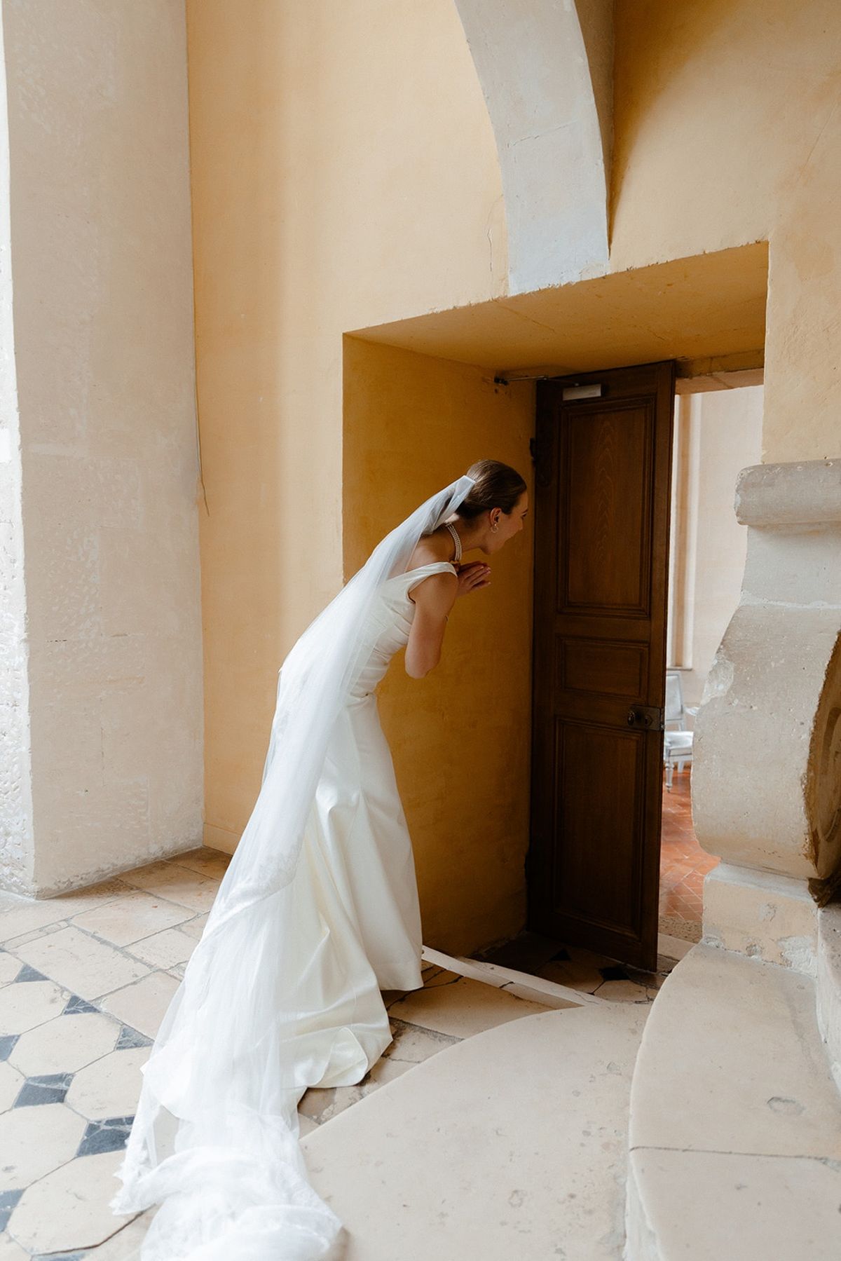 Bride leans against wooden door in sunlit ochre alcove with gown train pooling