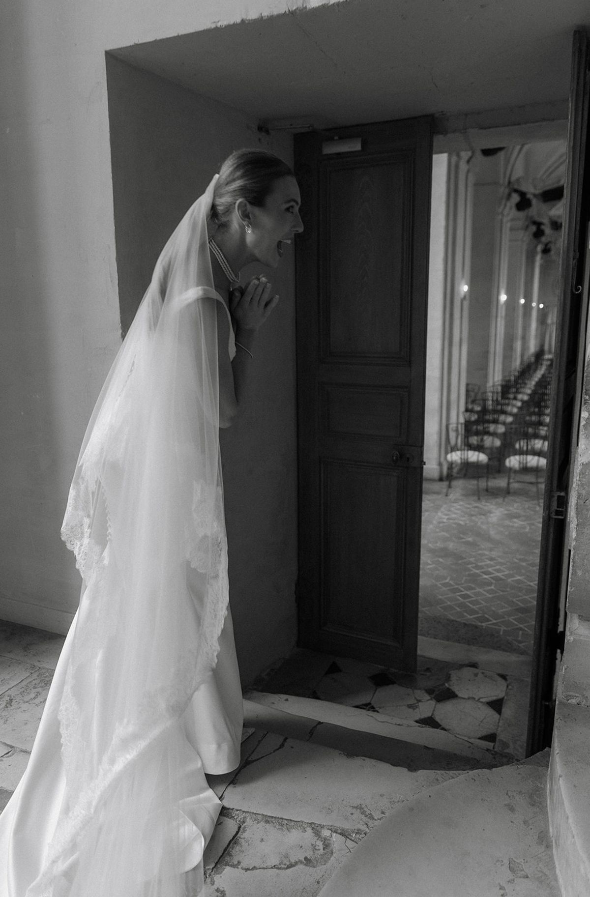 Bride peeks through wooden door toward ceremony hall with iron chairs visible
