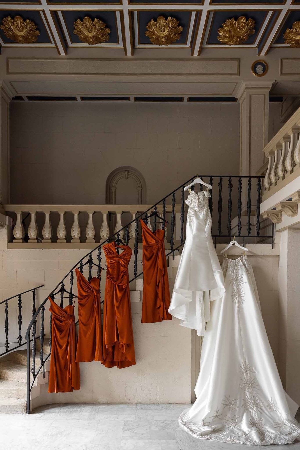Wedding dresses and burnt orange bridesmaid dresses hanging on grand staircase at Parc Mougins