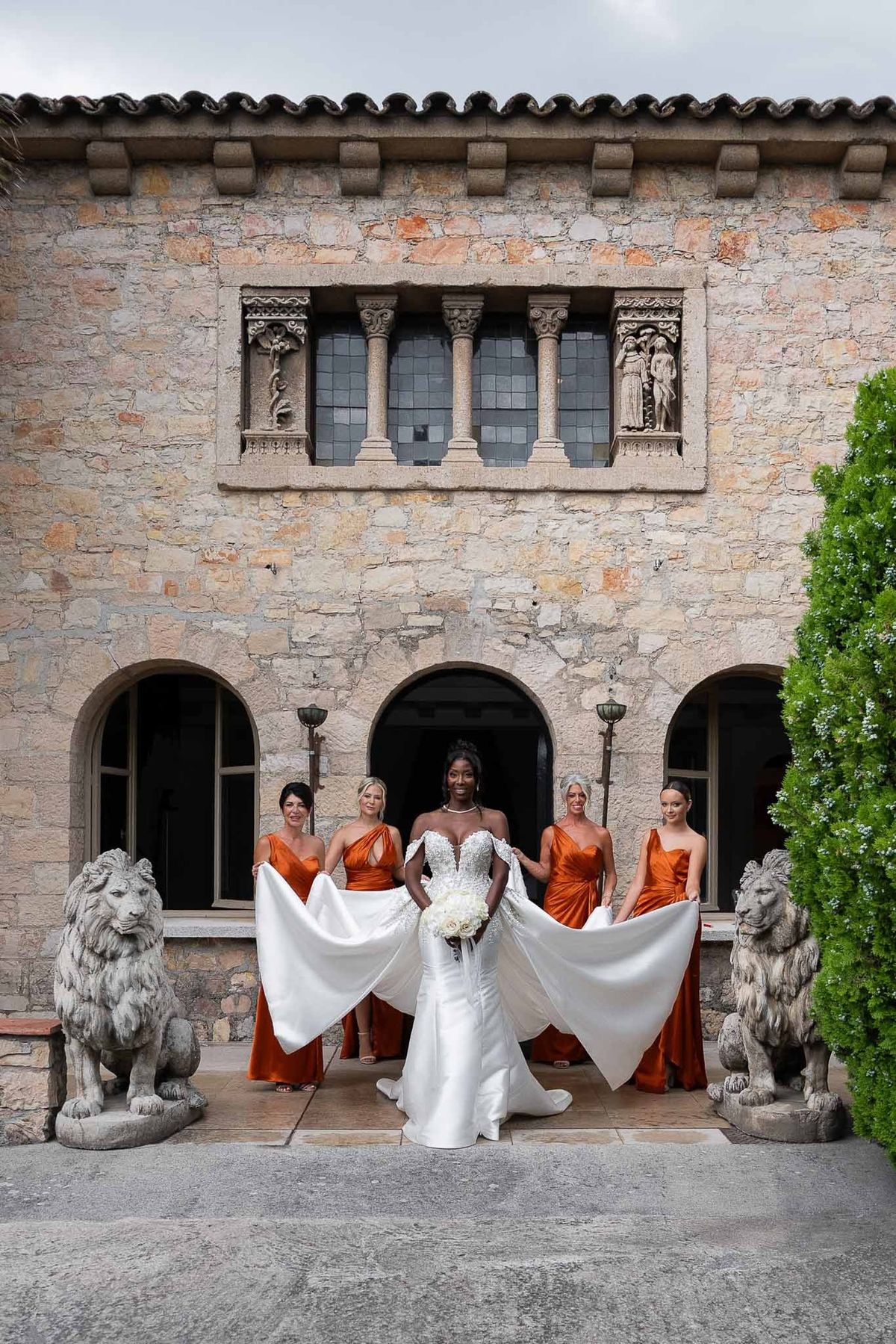 Bride and bridesmaids in burnt orange dresses at stone entrance with lion statues at Parc Mougins