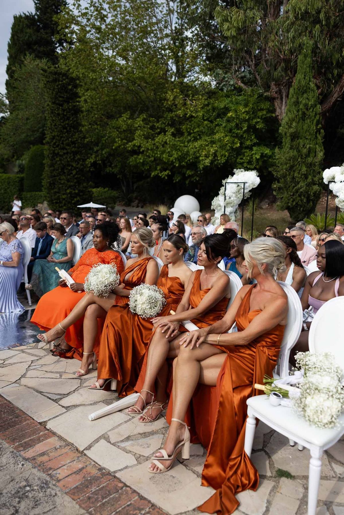 Bridesmaids in burnt orange dresses seated at poolside ceremony at Parc Mougins