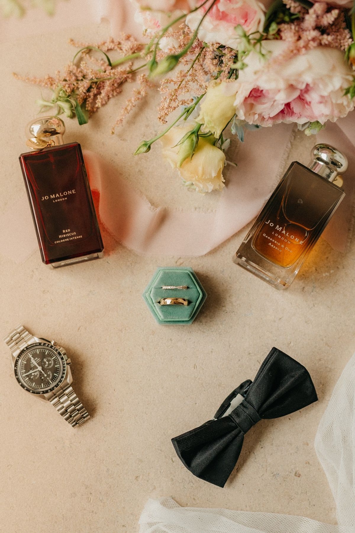 Overhead flat lay of wedding rings, perfumes, watch, bow tie and pastel florals on blush silk.