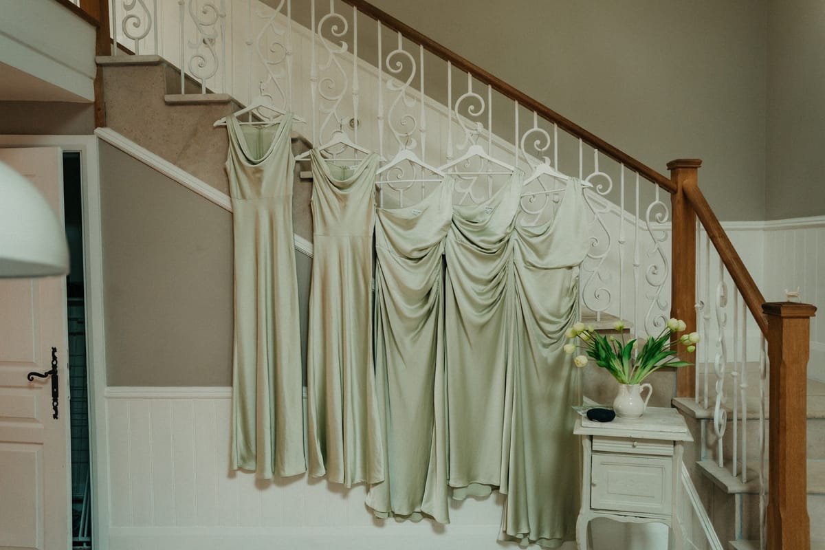 Five sage-green satin bridesmaid dresses on hangers along a white iron staircase.